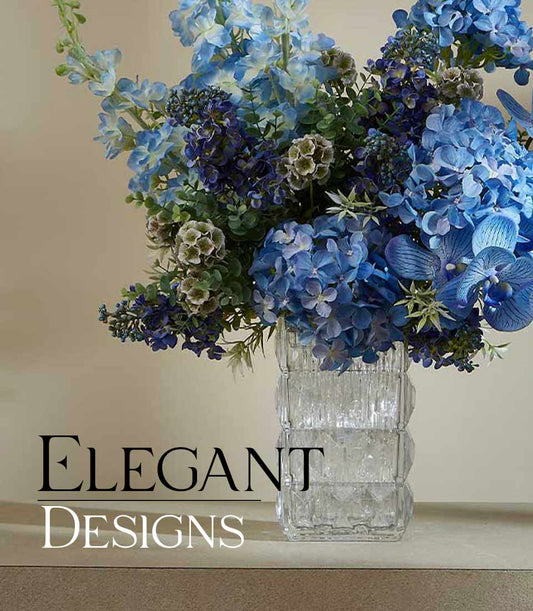 Blue and purple artificial flower arrangement in textured glass vase with delphiniums, hydrangeas, orchids, and foliage