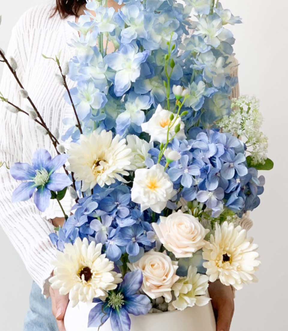 Blue and white artificial floral arrangement with hydrangeas, gerberas, and roses in white vase