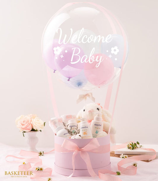 Hot air balloon baby gift set with pink box, organic care products, plush bunny, and pastel balloons on soft pink backdrop