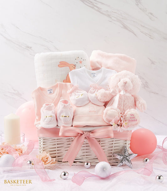 Delicate pink and white newborn gift basket with soft cotton clothing, plush teddy bear, and bunny-patterned blanket