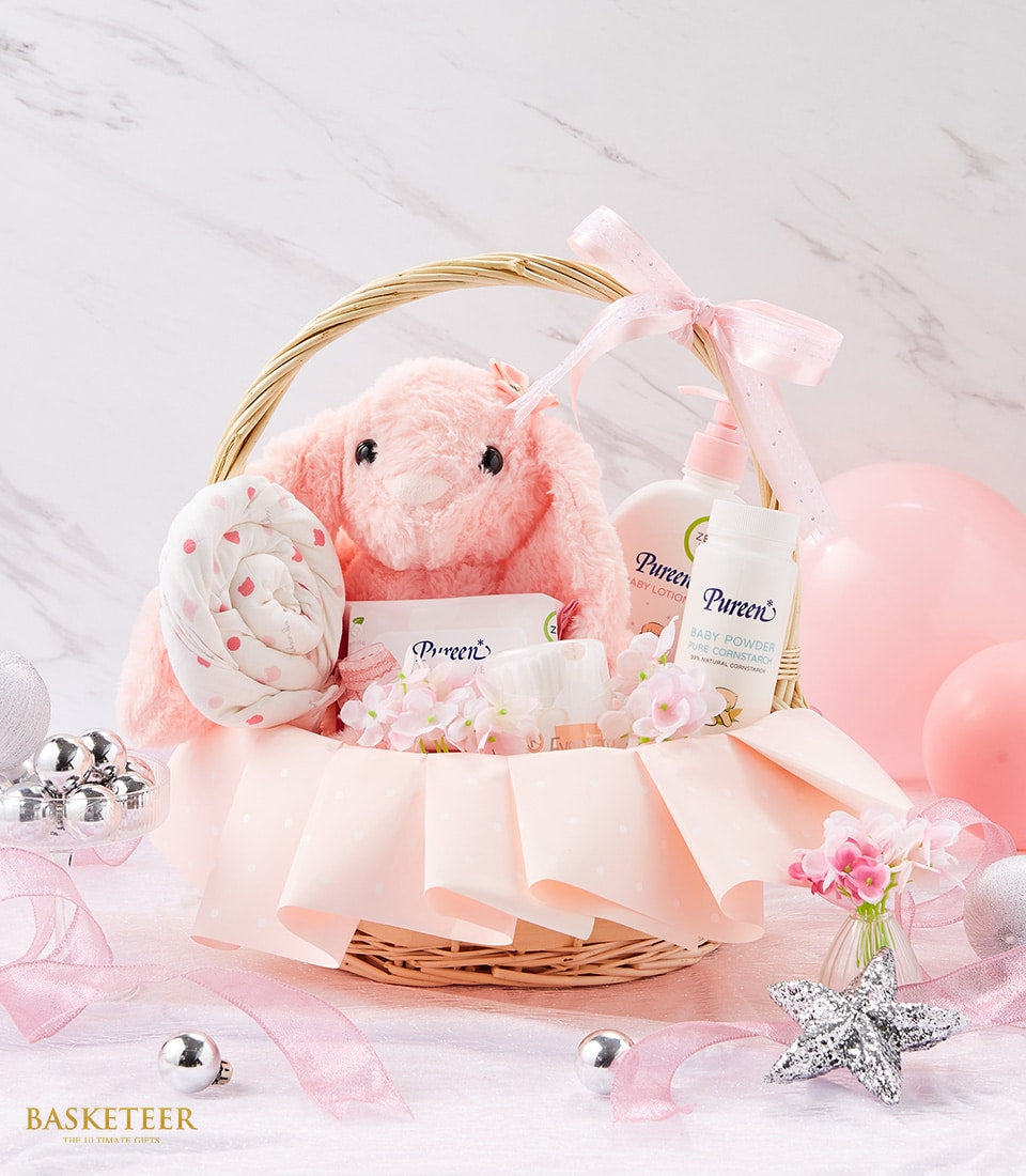 Wicker baby gift basket with pink polka dot blanket, plush bunny toy, and Pureen baby care products, light marble background