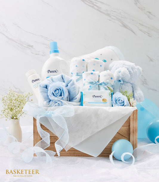 Blue and white newborn gift basket with baby essentials, plush teddy bear, and polka dot blankets in wooden crate