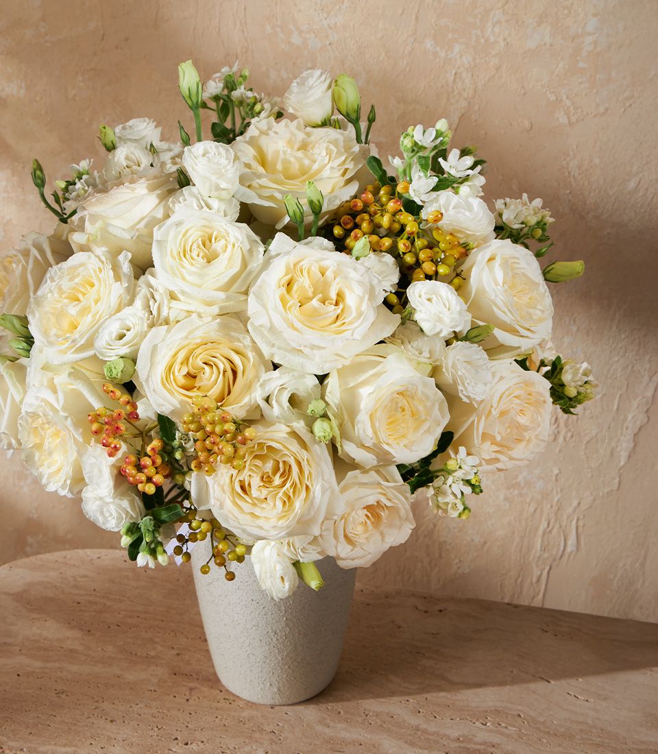 White and cream roses with yellow-center blooms in textured gray ceramic vase, accented with berries and greenery
