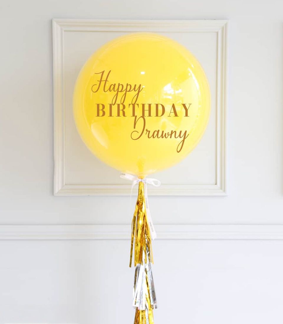 Vibrant yellow Happy Birthday balloon with gold and silver tassel, personalized message in brown script