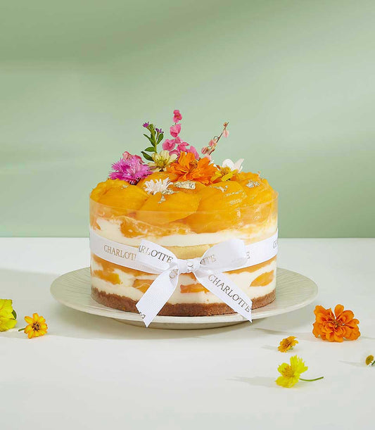 Layered mango cheesecake with fresh pink and orange flowers, gold leaf, and Charlotte Bakery ribbon on white plate