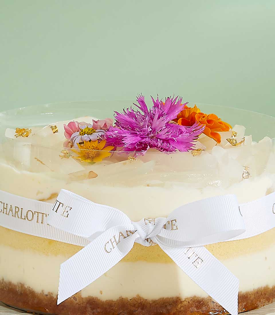 Creamy layered cheesecake with dark crumb base, fresh purple and orange flowers, and gold leaf in clear dome container