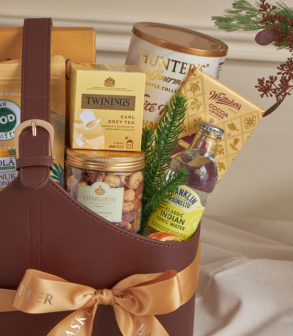 Premium brown leather gourmet gift basket with gold satin bow, filled with tea, chocolates, nuts, and festive greenery for