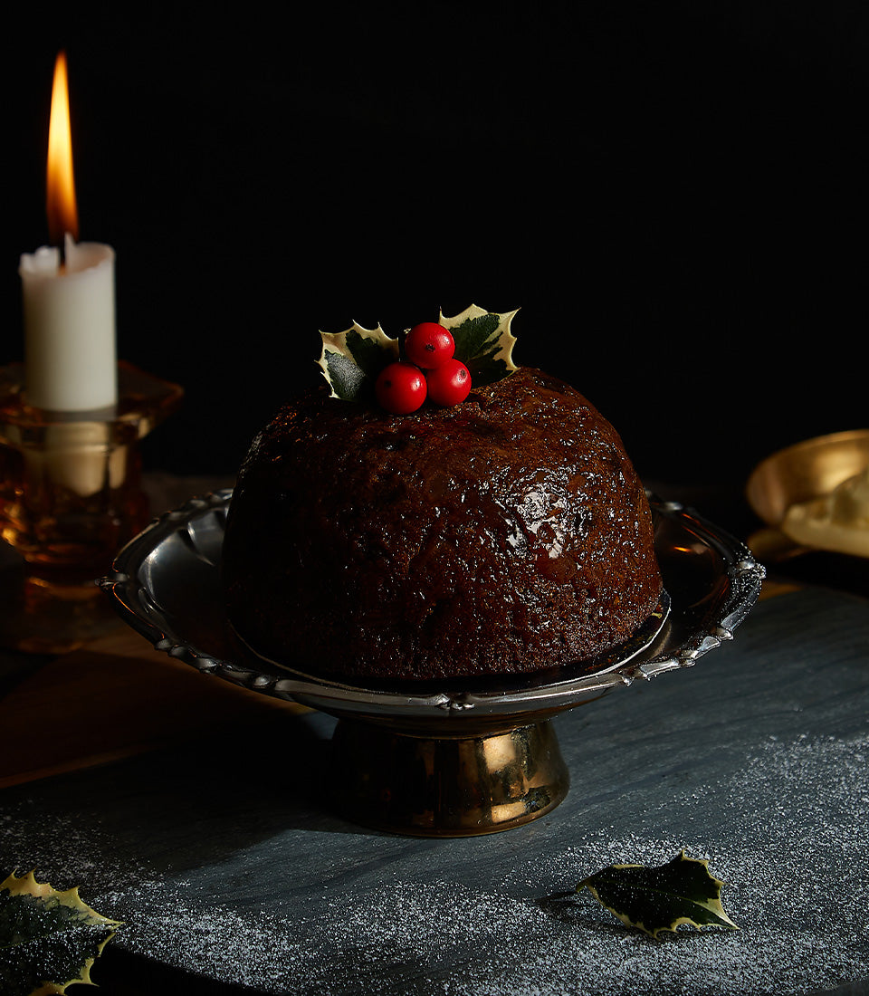 Dark traditional British Christmas pudding with holly berries and leaves on silver cake stand, festive holiday dessert