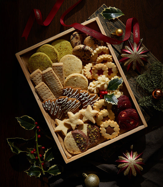 Wooden box of assorted Christmas cookies with star and fluted shapes, jam centers, and chocolate drizzles, surrounded by