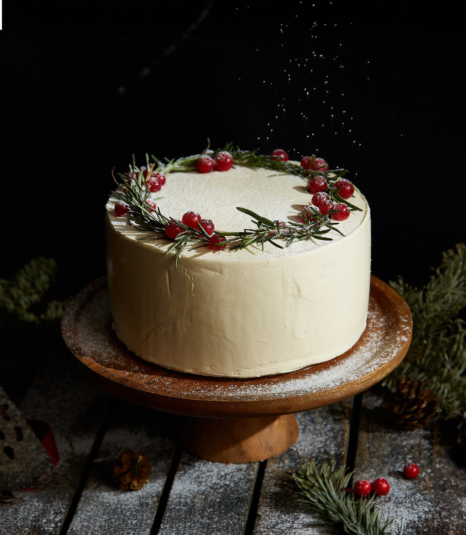 Festive white carrot cake with rosemary and cranberry wreath on wooden stand, powdered sugar snow for Christmas