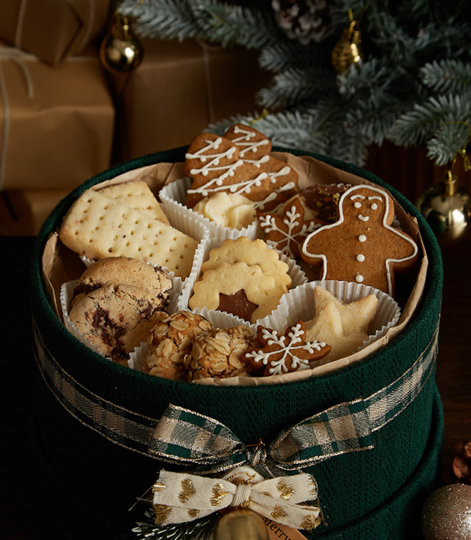 Dark green gift box of assorted Christmas cookies with gingerbread, trees, and stars in white paper cups, tied with plaid