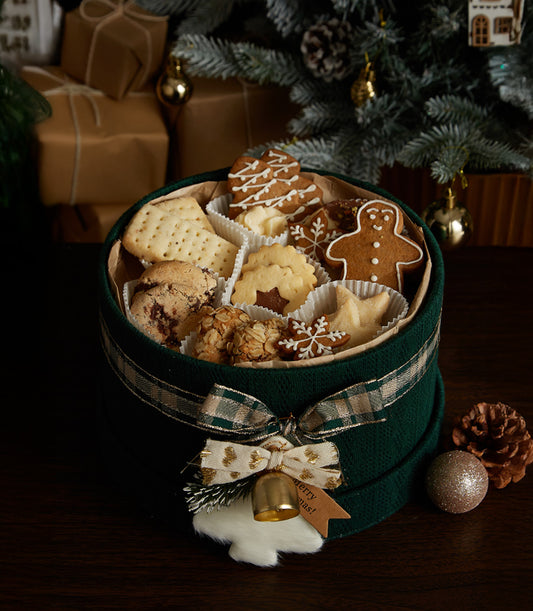 Festive green round gift box with Christmas cookies, gingerbread men, and crackers tied with plaid ribbon and gold bell