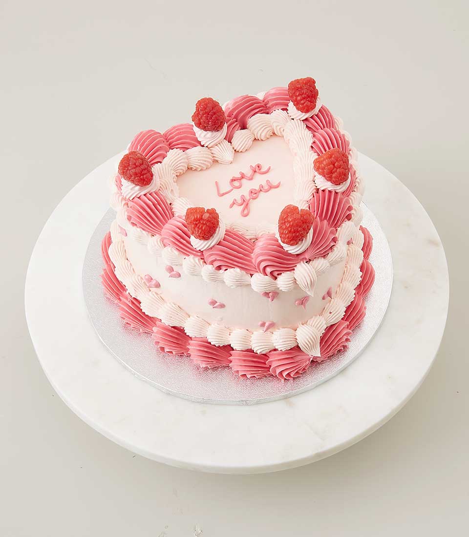 Heart-shaped vanilla cake with pink frosting, raspberries, and "Love you" message on marble stand