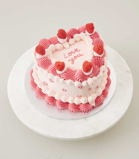 Heart-shaped vanilla cake with pink frosting, raspberries, and "Love you" message on marble stand