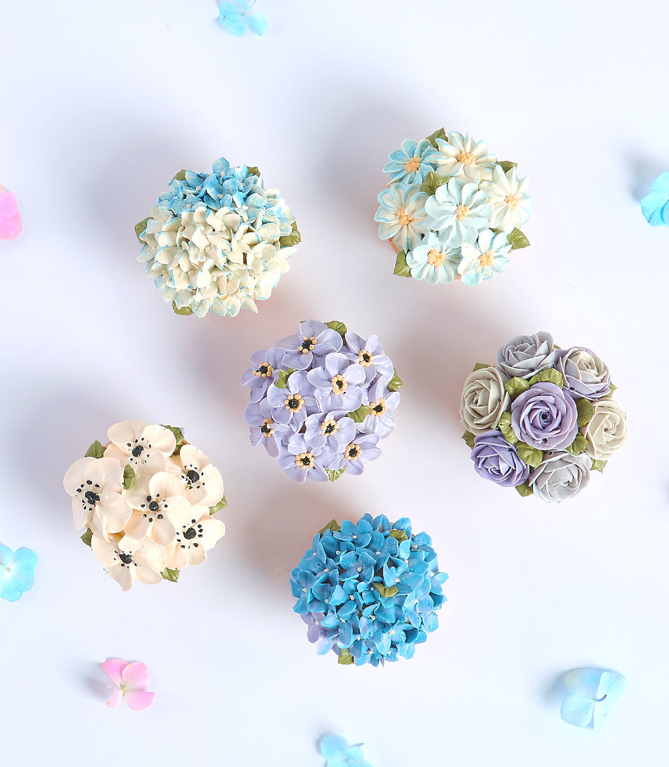 Six delicate floral cupcakes with buttercream hydrangeas, roses, and daisies in pastel blues and purples for Mother's Day