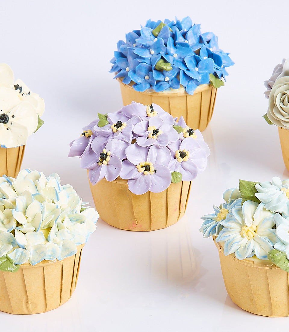 Six artisan cupcakes with hand-piped floral frosting in blue, purple, and white for Mother's Day