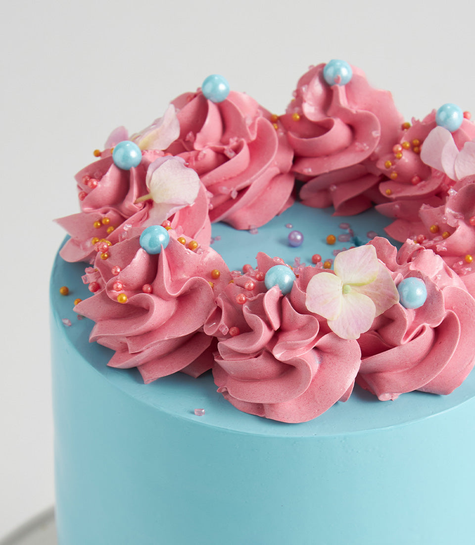 Pastel sky-blue celebration cake with pink frosting swirls, candies, and flower petals