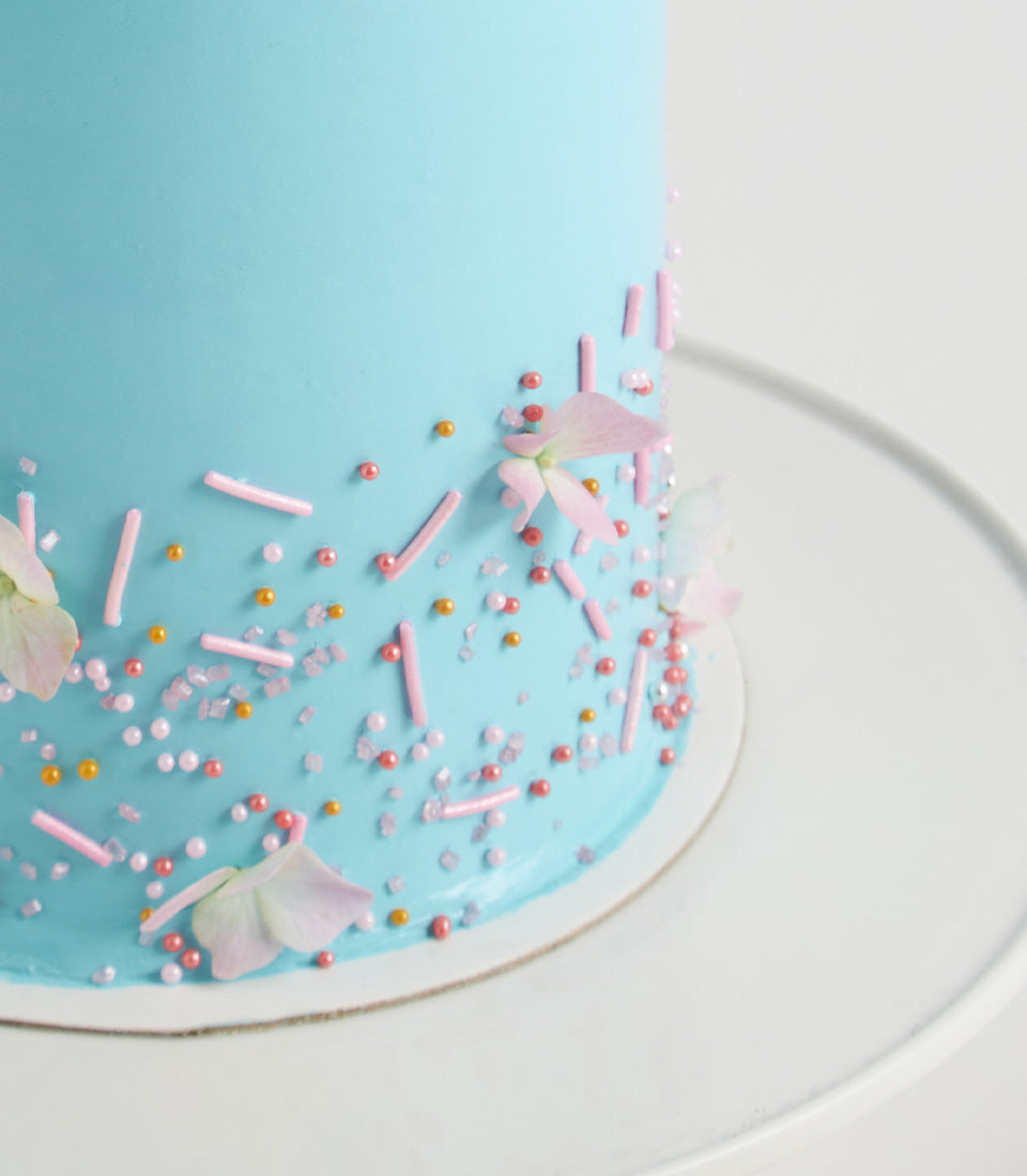 Light blue layer cake with pink and white edible flowers, pearl sprinkles, and sugar crystals for birthday celebrations