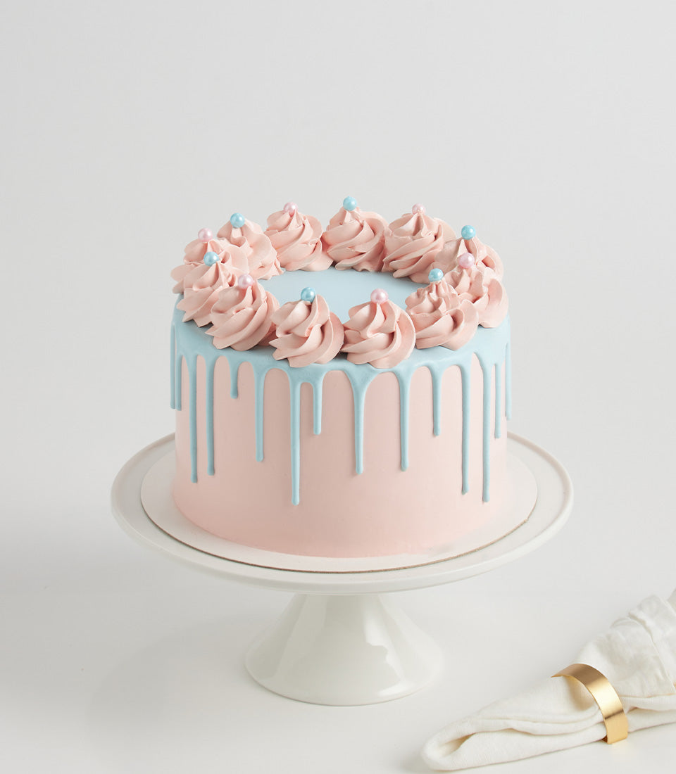 Pastel pink two-layer birthday cake with blue drip glaze and buttercream rosettes on white cake stand