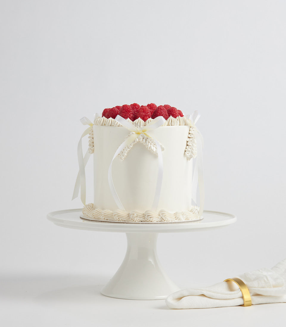 Elegant white buttercream birthday cake with fresh raspberries and satin ribbon on ceramic stand