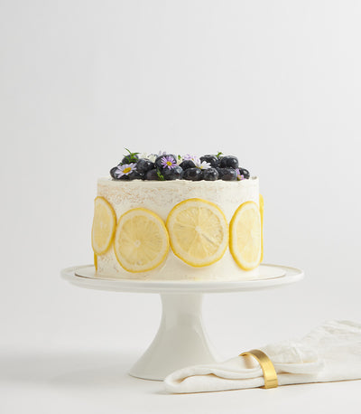 White layer cake with lemon slices and fresh blueberries, topped with purple and white flowers for birthday celebrations