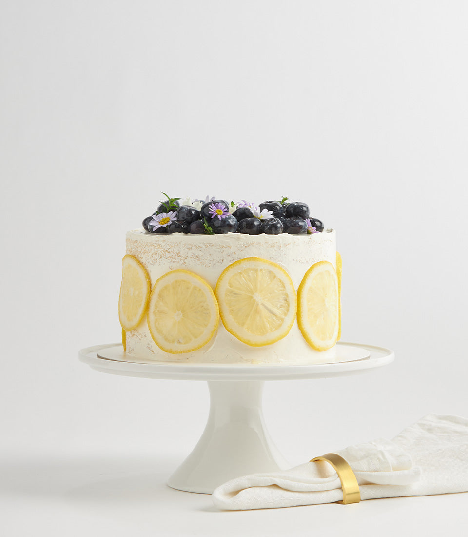 White layer cake with lemon slices and fresh blueberries, topped with purple and white flowers for birthday celebrations