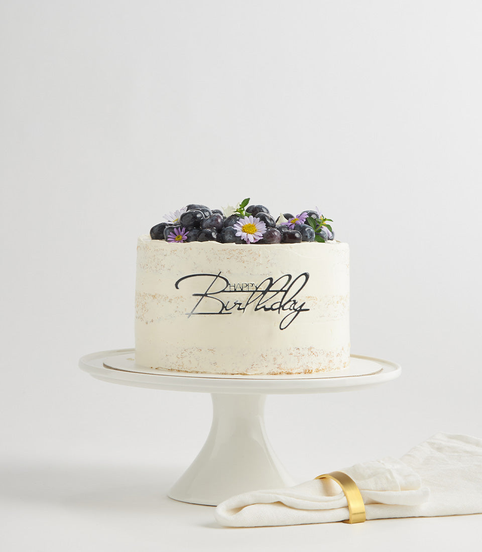 Semi-naked blueberry layer cake with purple grapes, flowers, and Happy Birthday topper on ceramic stand