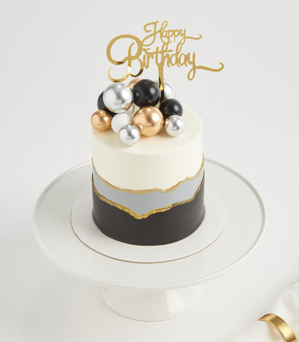 Luxe Black Forest two-tier birthday cake with white frosting, metallic gold accents, and gold Happy Birthday topper on white