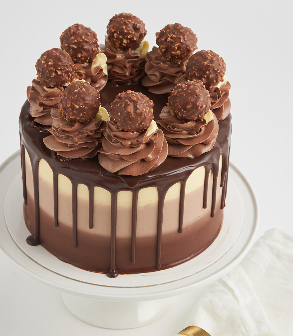 Ferrero Biscoff Bliss three-tier chocolate layer cake with ganache drip and truffle-topped swirls on white stand
