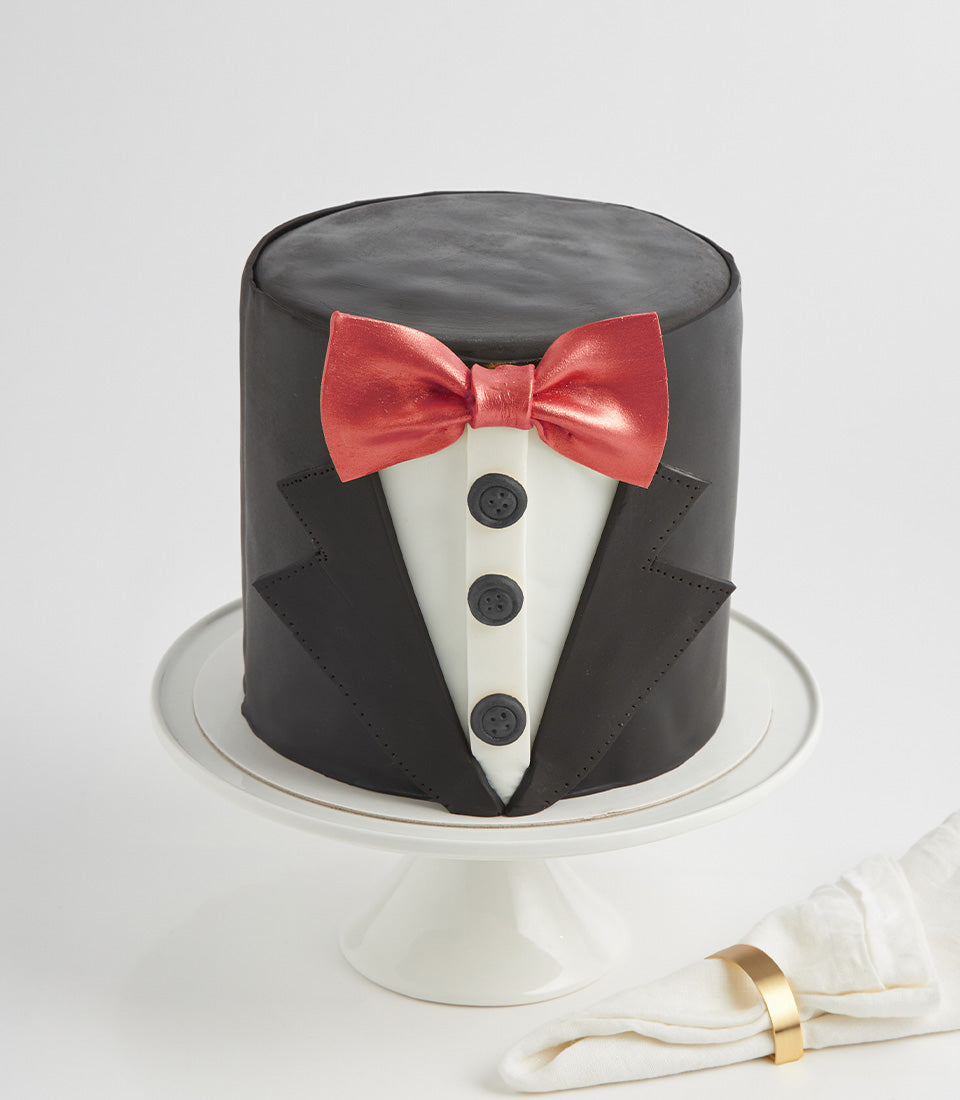 Black fondant tuxedo cake with white shirt front, red bow tie, and gold napkin ring detail