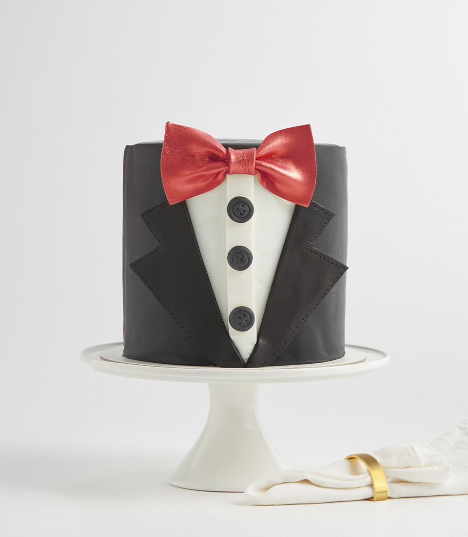 Black fondant tuxedo cake with red bow tie and white shirt front on pedestal stand