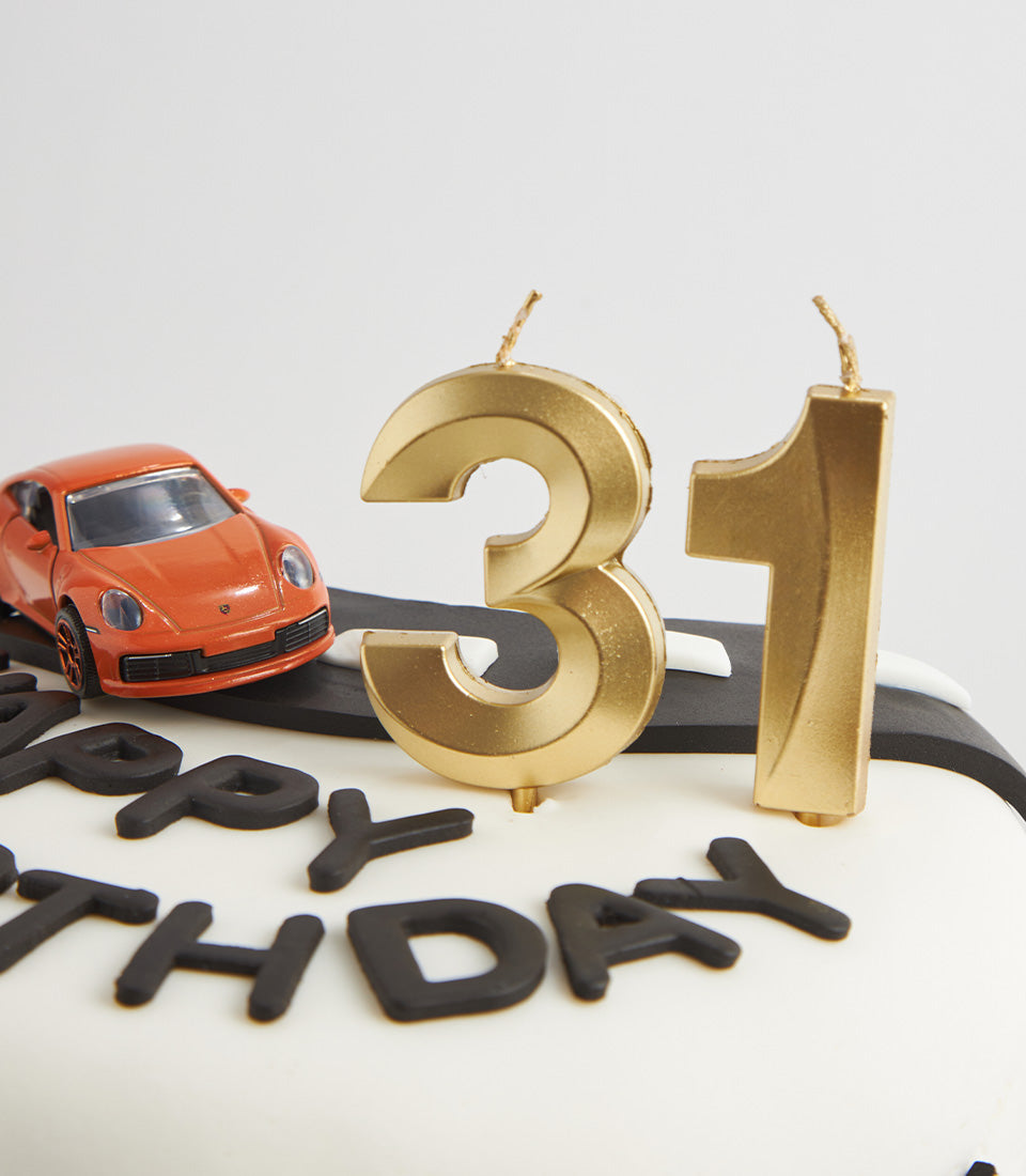 White fondant birthday cake with black road design, toy sports car, and golden numeral candles for 31st celebration