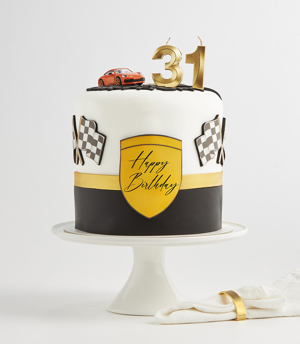 White and black two-tiered racing theme birthday cake with checkered flags, gold shield plaque, and toy sports car