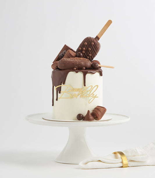 Chocolate drip birthday cake with chocolate bar toppings and gold Happy Birthday cake topper on white stand
