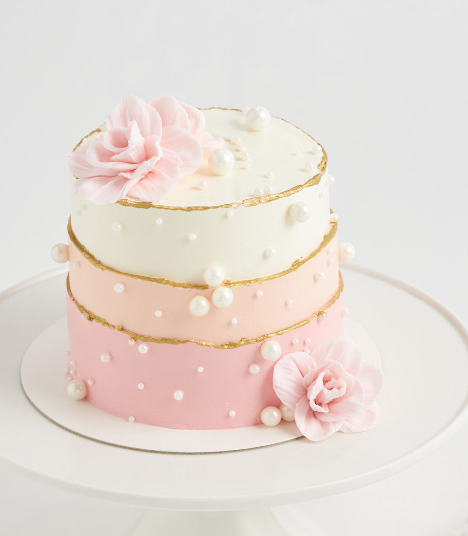 Three-tiered pearl birthday cake with ombre pink icing, edible gold trim, and sugar flowers on white stand