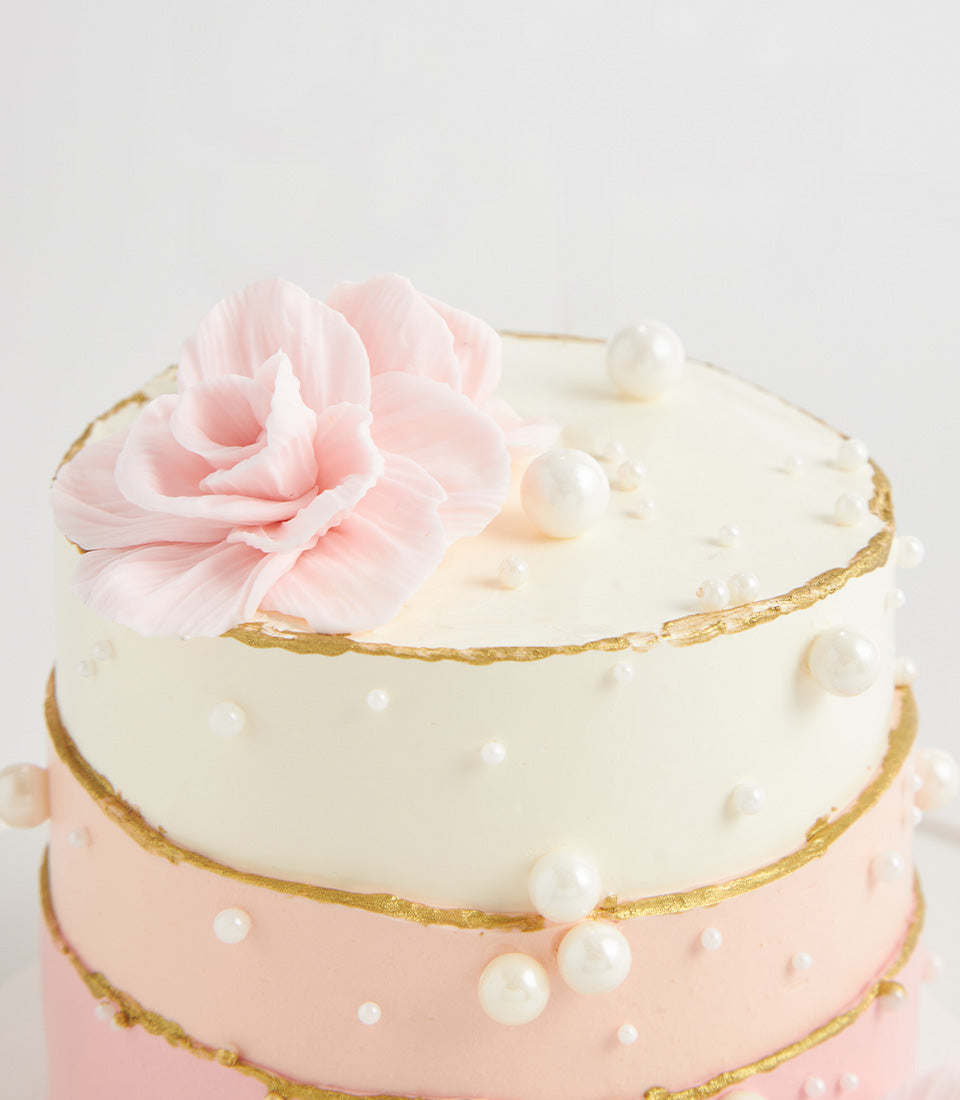 Elegant two-tiered birthday cake with cream and pink frosting, gold leaf details, and white edible pearls