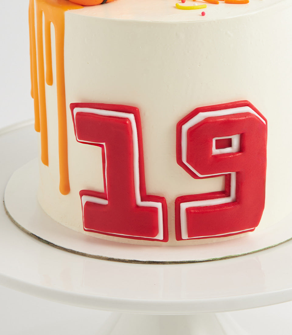 White frosted birthday cake with bold red "19" numerals and orange drip icing, topped with colorful sprinkles