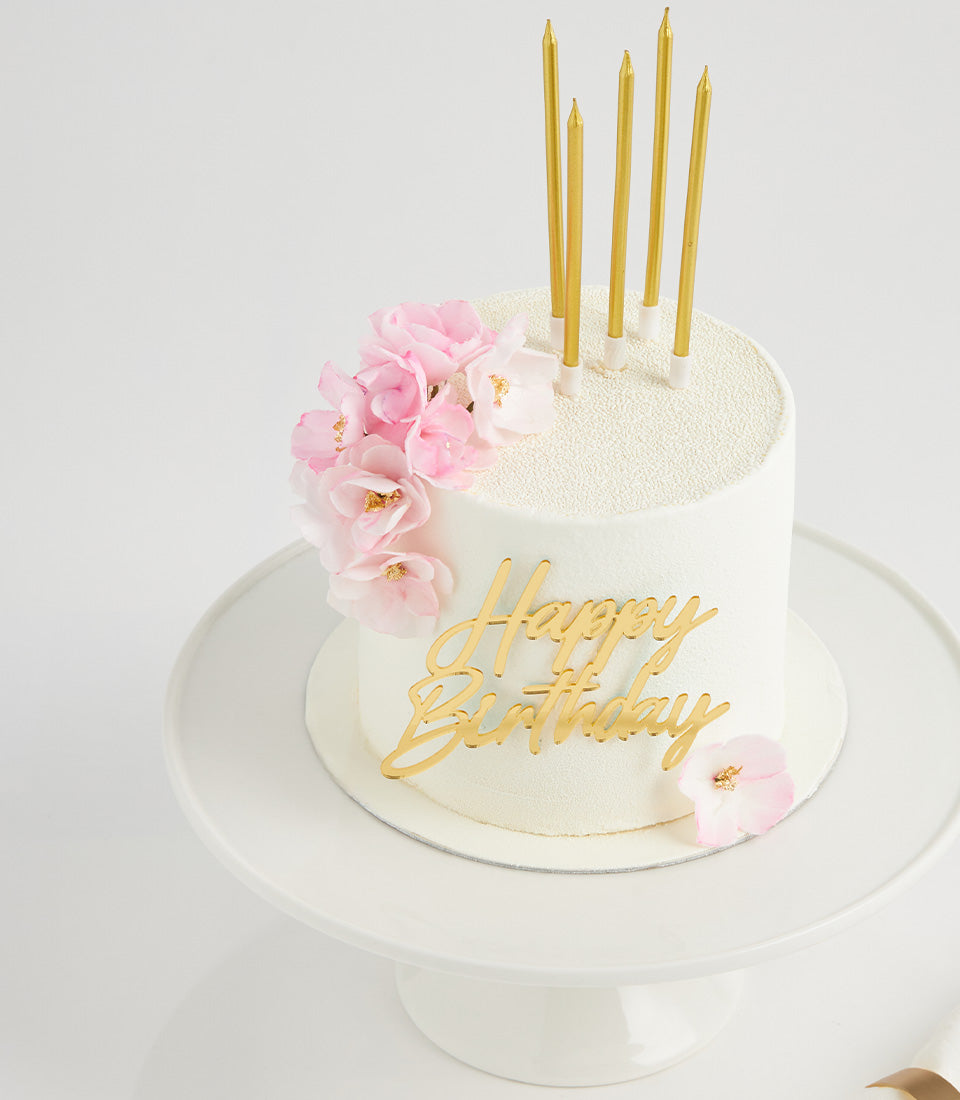 White birthday cake with pink flowers and gold accents on cake stand