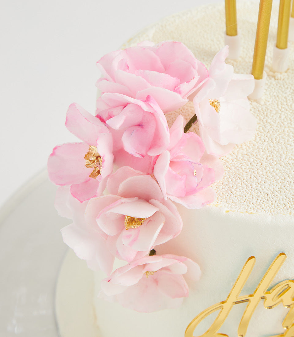 White frosted birthday cake with cascading pink sugar flowers and gold leaf accents