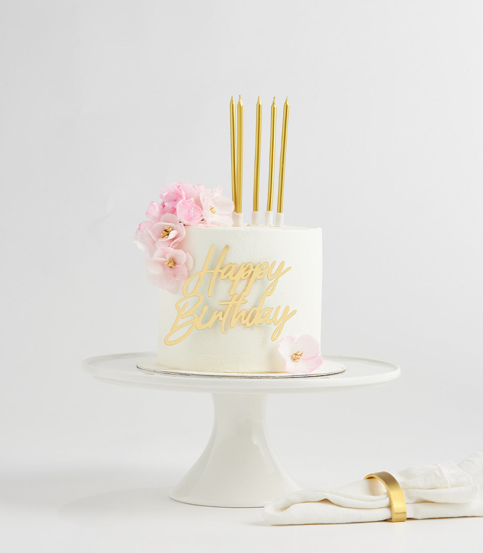 Elegant white birthday cake with pink flowers, gold candles and Happy Birthday topper on ceramic stand