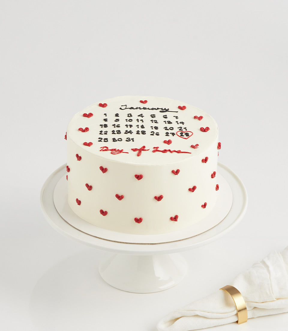 White round vanilla layer cake with black calendar design, red circled date, and Day of Love message with heart decorations