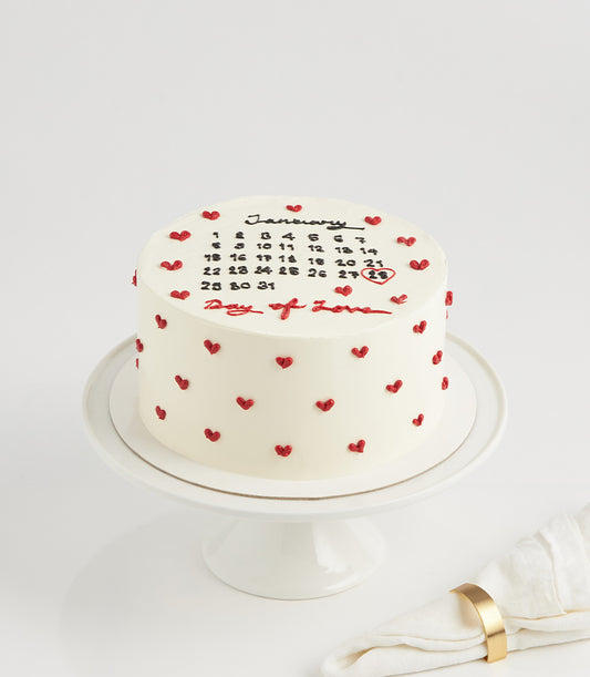 White round vanilla layer cake with black calendar design, red circled date, and Day of Love message with heart decorations