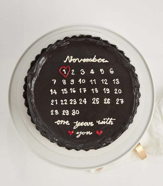 Dark chocolate anniversary calendar cake with November dates and "one year with you" message on ceramic stand