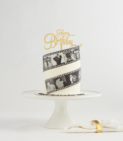 White two-tier birthday cake with black and white edible film strip photos and gold Happy Birthday topper