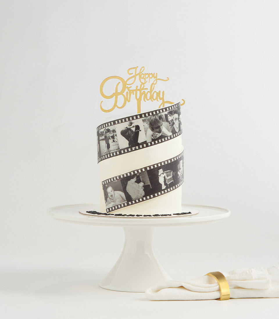 White two-tier birthday cake with black and white edible film strip photos and gold Happy Birthday topper