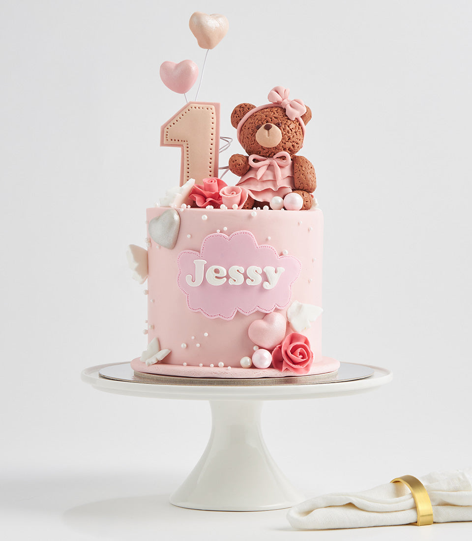 Light pink first birthday cake with teddy bear, hearts, and personalized name plaque on ceramic stand