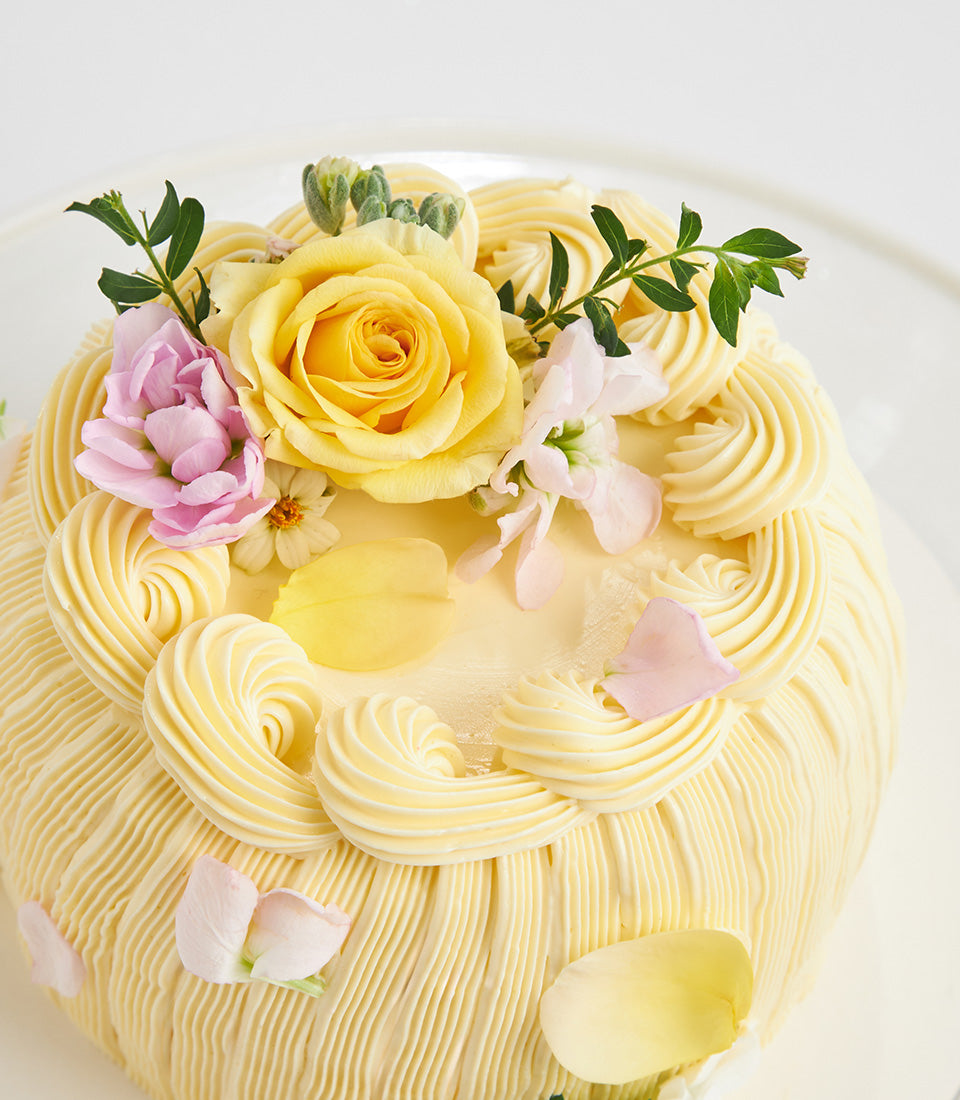 Pale yellow layer cake with fresh flowers and pink blossoms on white stand, artisan birthday cake