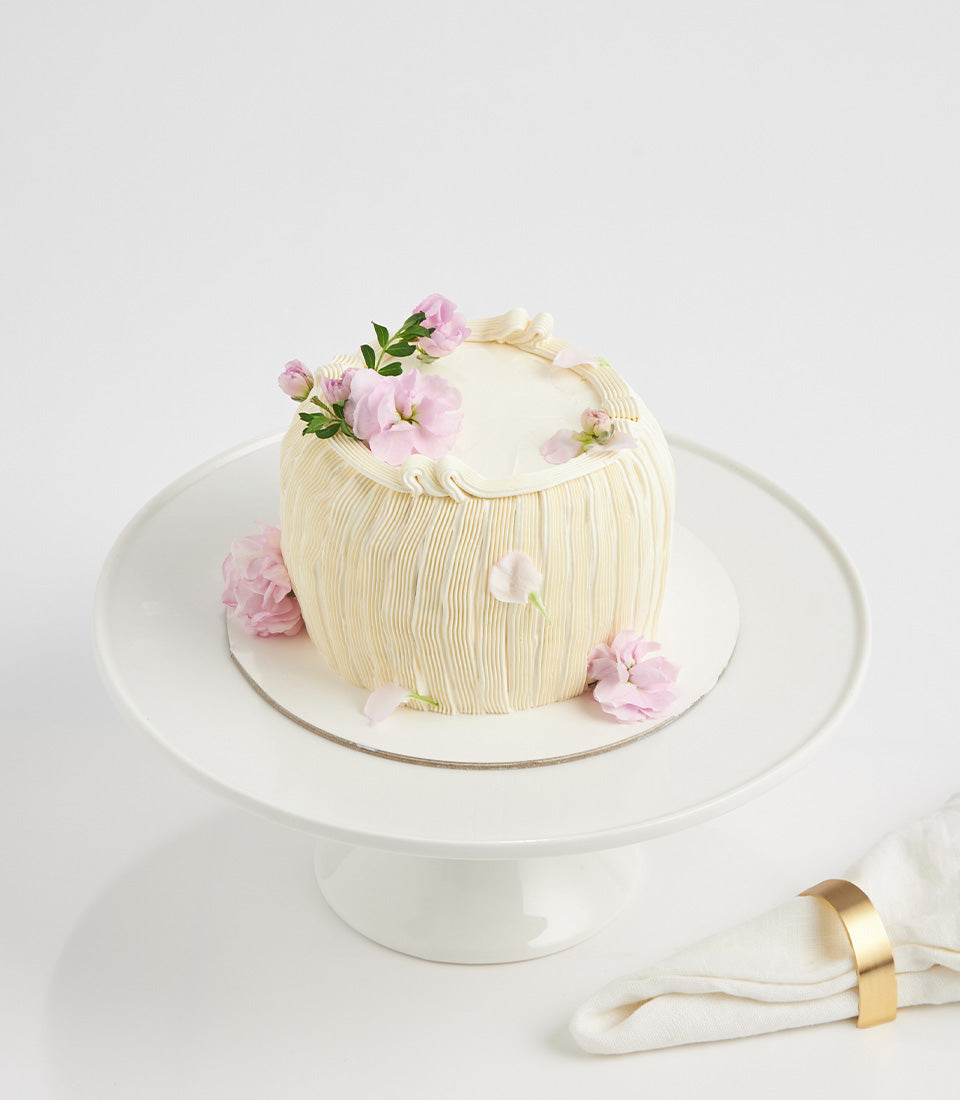 Lemon Cream Blossom cake with pleated frosting, fresh pink flowers, and gold-trimmed napkin on white stand
