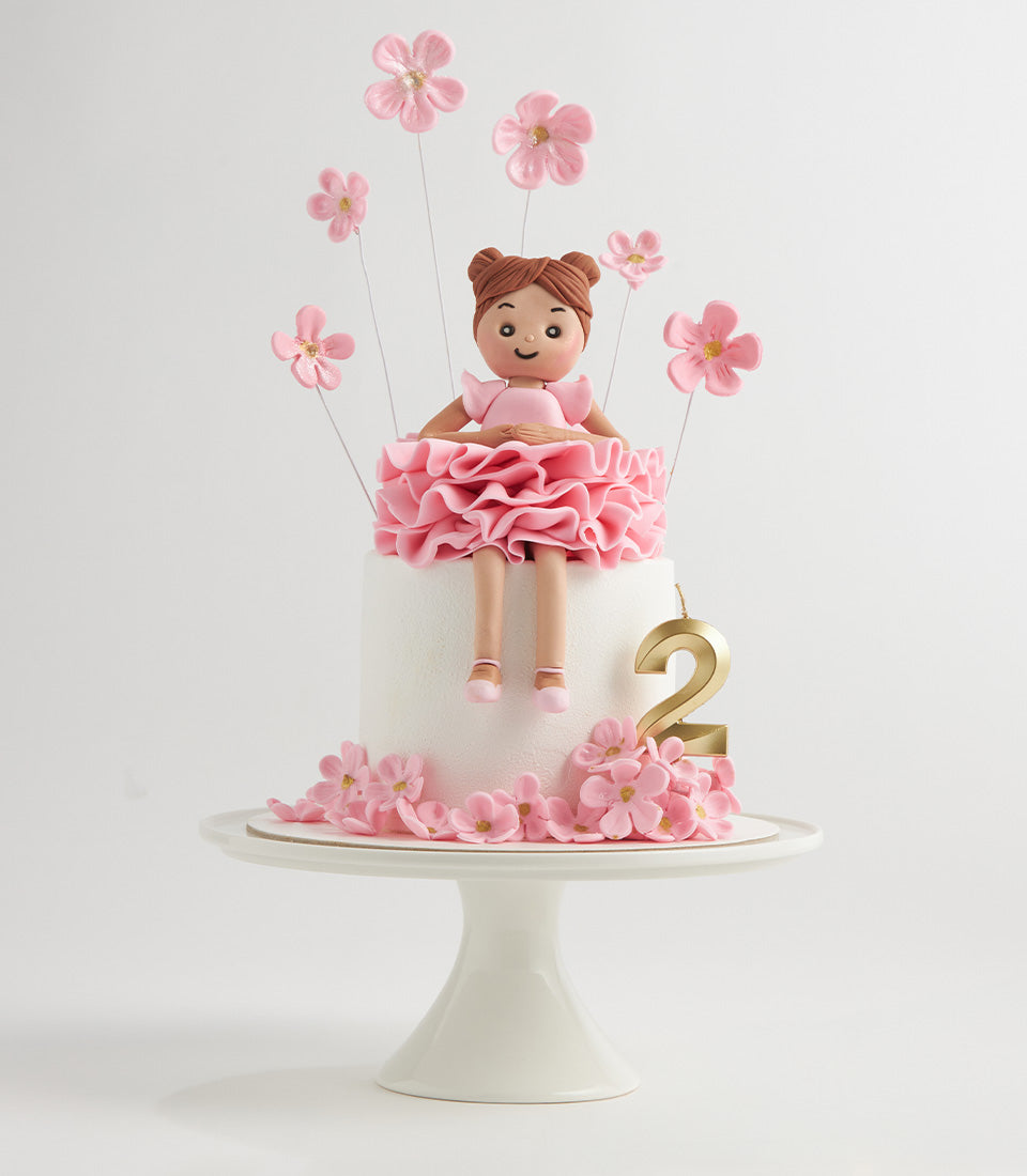 Two-tier white birthday cake with pink fondant ballerina doll, sugar flowers, and gold number candle for second birthday