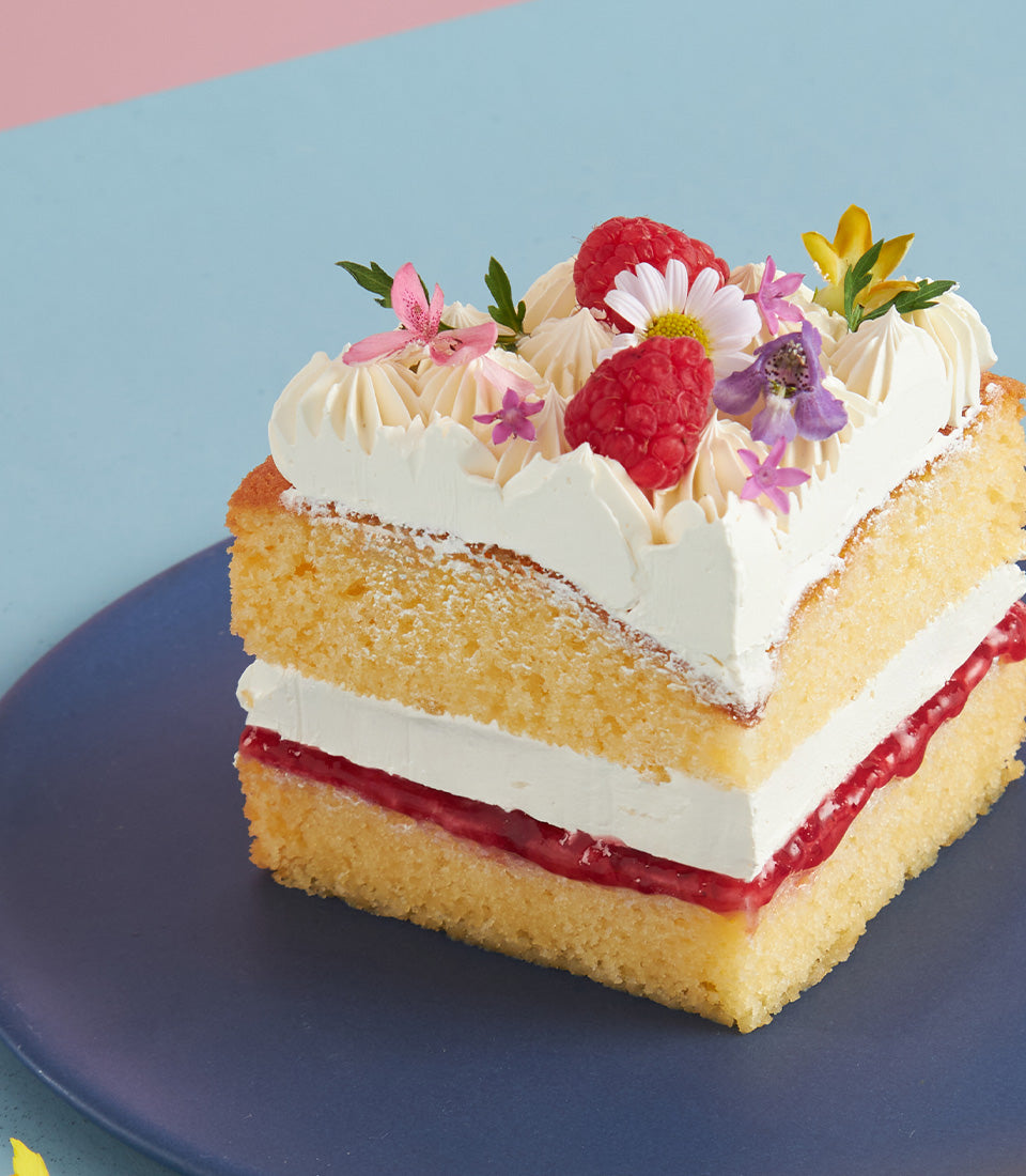 Raspberry buttercream sponge cake slice with fresh berries and edible flowers on dark blue plate